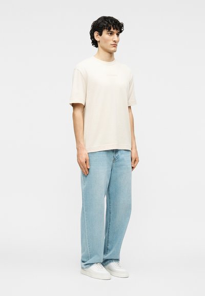 Νεαρός άνδρας με σγουρά μαλλιά στέκεται, φορώντας ένα απλό υπερμεγέθες μπλουζάκι σε μπεζ χρώμα, ανοιχτόχρωμα τζιν με φαρδιά μπατζάκια και λευκά αθλητικά παπούτσια.