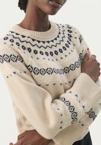 Pull en tricot beige avec des motifs géométriques bleu marine et blancs, doté d'un col rond et de poignets côtelés. Texture douce, coupe décontractée.