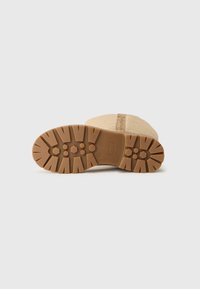 Suela de bota de ante color beige con una suela de goma texturizada. Presenta patrones circulares y hexagonales para mejorar la tracción. Costura visible cerca del borde superior.