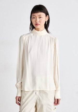 Long-sleeve cream blouse with a high neckline, gathered shoulder seams, and cuffed sleeves, made from a smooth, lightweight fabric.