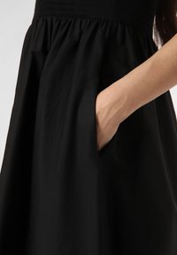 Schwarzes Kleid mit gerafftem Oberteil und flarem Rock sowie seitlichen Taschen. Glatte Textur, leichter Stoff und einfarbig.