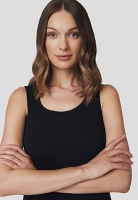 Czarny bezrękawnik o okrągłym dekolcie, wykonany z gładkiego materiału. Modelka ma długie, falowane włosy i krzyżuje ramiona.