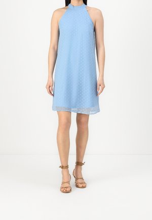 Nainen, joka pukeutunut hihattomaan vaaleansiniseen polvipituiseen mekkoon, jossa on hienovarainen pilkkukuvio ja ruskeat remmikkäät korkokengät.