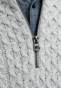 Hellgrauer gestrickter Pullover mit einem strukturierten Kabelmuster, einem Frontreißverschluss und einem blauen Hemd, das darunter sichtbar ist.