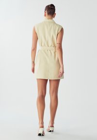 Abito senza maniche di un giallo chiaro con vita cintrata, dotato di colletto e una trama liscia. Luce corta, abbinato a tacchi bianchi.