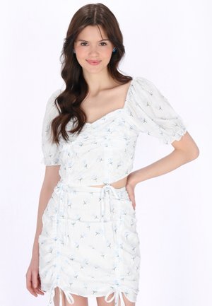 Młoda kobieta uśmiechająca się z długimi brązowymi włosami, ubraną w biały kwiatowy marszczony krótki top i dopasowaną spódnicę, stojąca na jednolitym białym tle.