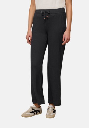 Persoon draagt losse zwarte gestructureerde broek met trekkoord in de taille en crème sneakers met zwarte accenten, staand op een witte achtergrond.