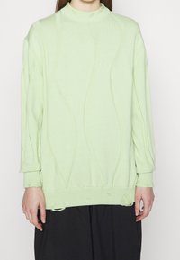 Ljust grönt stickat tröja med texturerade vågmönster, hög krage och något fransiga fållar och ärmslut. Långa ärmar med en lös passform.