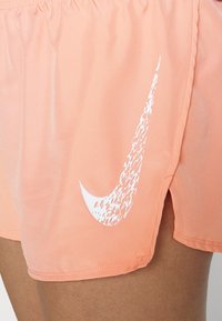 Urheilushortsit vaalean persikan värisestä kankaasta, joka on sileä rakenne. Sivussa on suuri valkoinen Nike-swoosh-logo ja joustava vyötärö. 