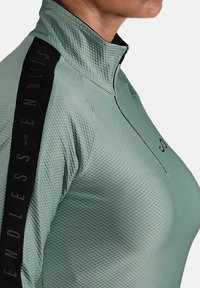 Lichtgroene sporttop met een textuurpatroon, hoge kraag en zwarte verticale streep met "ENDLESS" in vetgedrukte letters.
