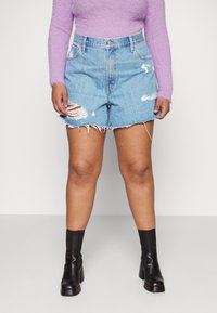 Shorts de mezclilla en azul claro con bordes deshilachados y detalles desgastados. Combinados con una blusa de manga larga morada de textura suave y botas negras de tobillo.