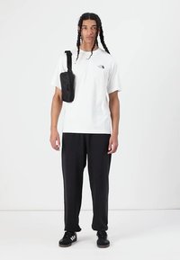 White cotton t-shirt with a logo, black trousers, and black sneakers. A small black bag is worn across the body. Simple, relaxed fit.