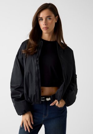 Giovane donna con capelli castani lunghi indossa un top nero corto, una giacca nera oversize con zip, jeans blu scuro e una cintura nera con conchiglie bianche.