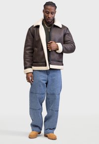 Bruine leren jas met witte bontkraag en -manchetten, gecombineerd met blauwe baggy jeans en beige schoenen, voorzien van een rits en zijzakken.