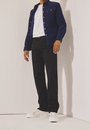 Personne portant un t-shirt blanc, une chemise bleu foncé à boutons, un pantalon noir et des baskets blanches, debout contre un mur clair et uni.