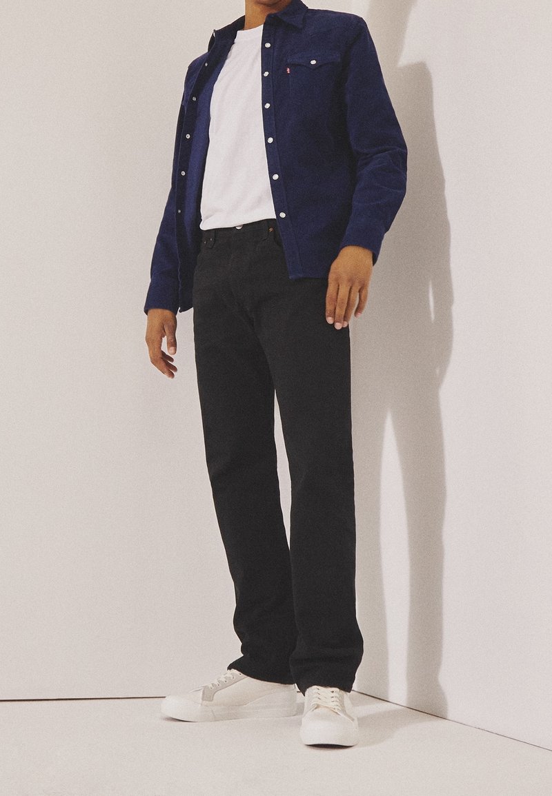 Personne portant un t-shirt blanc, une chemise bleu foncé à boutons, un pantalon noir et des baskets blanches, debout contre un mur clair et uni.