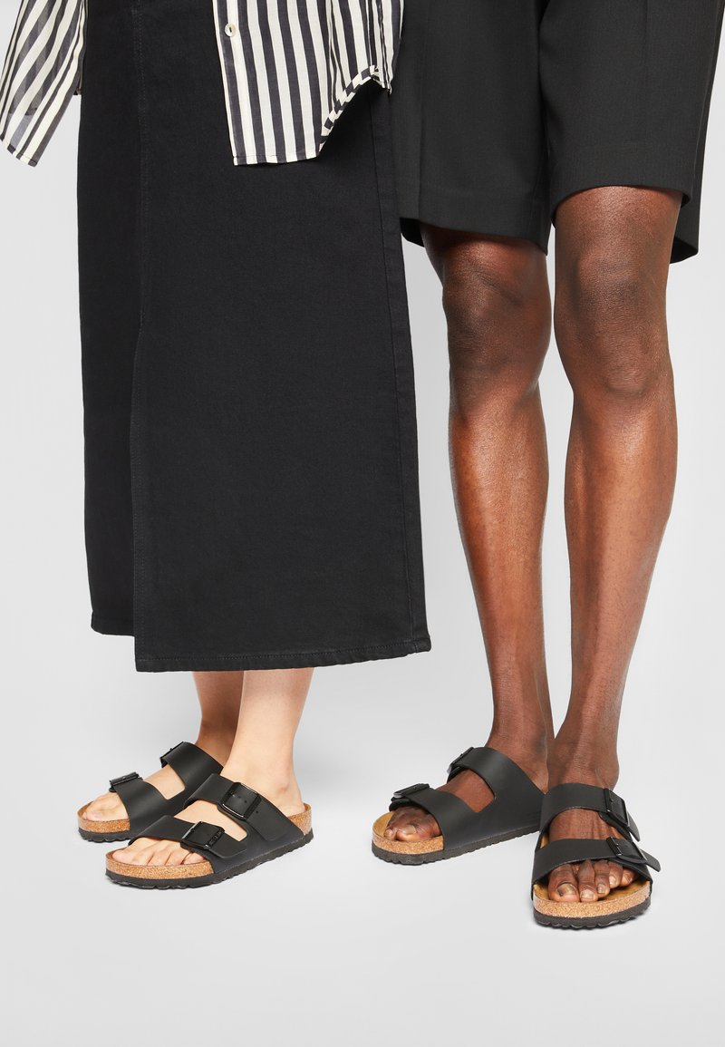 Black sandals with double buckle design and cork footbed. Textured rubber sole. One pair worn by a person with light skin, the other by a person with dark skin.