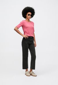 Γυναίκα με αφρικανικό χτένισμα, φορώντας γυαλιά ηλίου, ροζ κοντομάνικη μπλούζα, μαύρα ψηλοκάβαλα τζιν και μπεζ αθλητικά παπούτσια, στέκεται με το χέρι στη μέση.