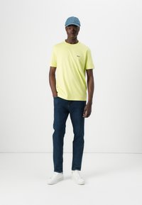 Lys gul T-skjorte med kort arm og en liten logo. Bæres med mørkeblå jeans og hvite joggesko, komplementert av en lyseblå caps.