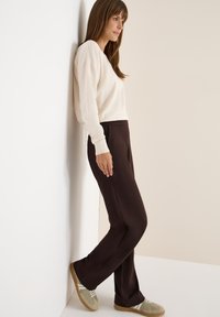 Ljusbeige tröja, bruna byxor och grönbruna sneakers. Modellen lutar sig mot en vägg och visar upp en avslappnad, casual outfit.