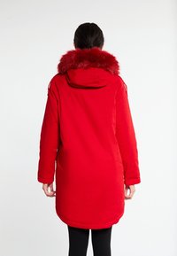 Parka rouge avec une capuche bordée de fourrure, manches longues et ourlet droit. Confectionnée dans un tissu lisse avec un rembourrage pour plus de chaleur.