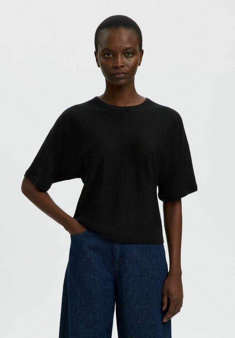 Femme aux cheveux courts portant un haut noir ample à manches courtes et un large pantalon en jean bleu, debout avec une main dans la poche.