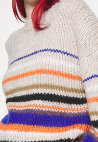 Pull en maille à col rond, présentant des rayures horizontales en orange, noir, bleu, beige et blanc, avec une finition douce et texturée.