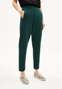 Persoon in donkergroene, taps toelopende broek met zijzakken en witte sneakers met zebra-print op een witte achtergrond.