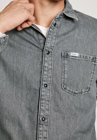 Graues Denimhemd mit Button-Down-Kragen, schwarzen Knöpfen und einer linken Brusttasche mit weißem Etikett. Strukturierter Stoff mit lässigem Design.