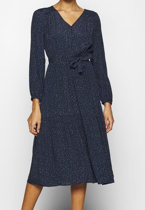 Marineblauwe midi wikkeljurk met lange mouwen en kleine witte stippen, gestrikt in de taille met een stoffen riem.