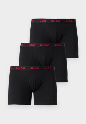 Jeune homme debout pieds nus portant un boxer noir avec une ceinture rouge portant le logo Hugo sur un fond clair uni.