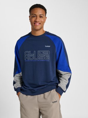 Mand smilende, iført en marineblå, kongeblå og grå sweatshirt med teksten "CLUB HOUSE" og beige bukser med mærkelogoer. Hænder i lommerne.