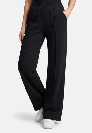 Zwarte wijde broek met elastische tailleband en voorste naden, gedragen met witte sneakers, één hand in de zak, tegen een effen achtergrond.