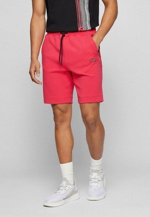 Homme portant un short décontracté rouge avec un cordon noir, des chaussettes blanches, des baskets blanches et un t-shirt noir avec un motif rayé sur fond gris.