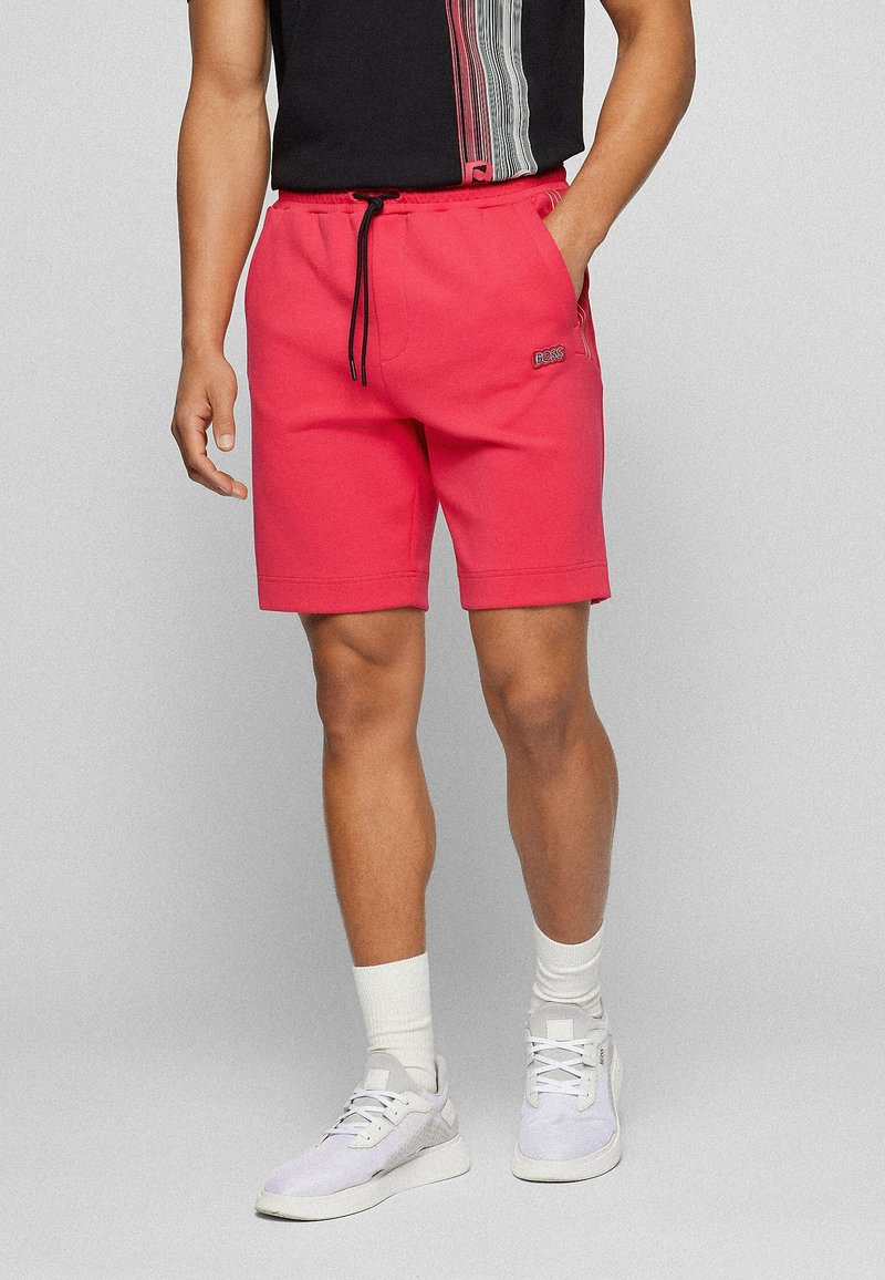 Homme portant un short décontracté rouge avec un cordon noir, des chaussettes blanches, des baskets blanches et un t-shirt noir avec un motif rayé sur fond gris.