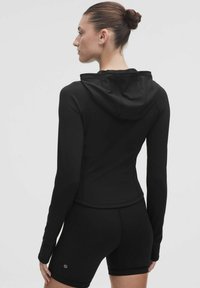 Jachetă sport neagră cu un design ajustat, glugă și mâneci lungi, asociată cu pantaloni scurți negri asortați. Textură netedă, branding minim.