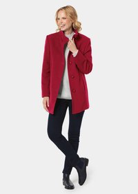 Roter Wollmantel mit hohem Kragen und fünf Knöpfen, kombiniert mit einem hellgrauen Pullover, dunklen Jeans und schwarzen Stiefeletten. Einfache Hintergrund.