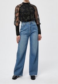 Blouse noire transparente avec des motifs floraux brodés, col haut et manches longues, associée à un jean en denim bleu à taille haute et coupe large.