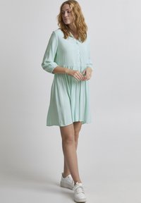 Rochie verde menta dintr-un material ușor, cu guler, cu închidere cu nasturi pe față și talie strânsă. Poartă adidași albi.
