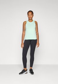 Een vrouw staat zelfverzekerd in een lichtgroene sporttop, zwarte leggings en sportieve schoenen, glimlachend tegen een effen achtergrond.
