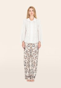 Blusa bianca a maniche lunghe con dettagli ricamati, abbinata a pantaloni larghi a stampa marrone e crema. Sandali semplici completano il look.