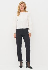 Witte cropped blouse met verticale plooitjes en lange pofmouwen, gecombineerd met zwarte slim-fit broek en zwarte schoenen met blokhak.