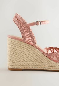 Sandale compensée tressée rose avec bride à la cheville et talon plateforme en jute tissée sur fond neutre.