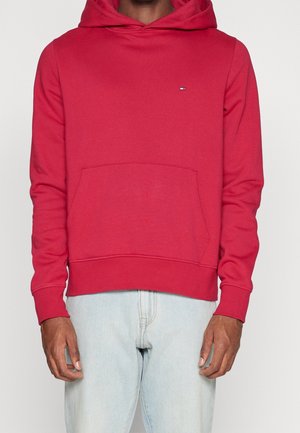 Homme portant un sweat à capuche rouge avec une poche avant et un petit logo sur la poitrine, associé à un jean bleu clair.