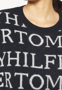 Schwarzer Strickpullover mit großen weißen Schriftzügen auf dem Stoff. Gerippter Rundhalsausschnitt und weiche Textur mit leichten Pillingstellen.