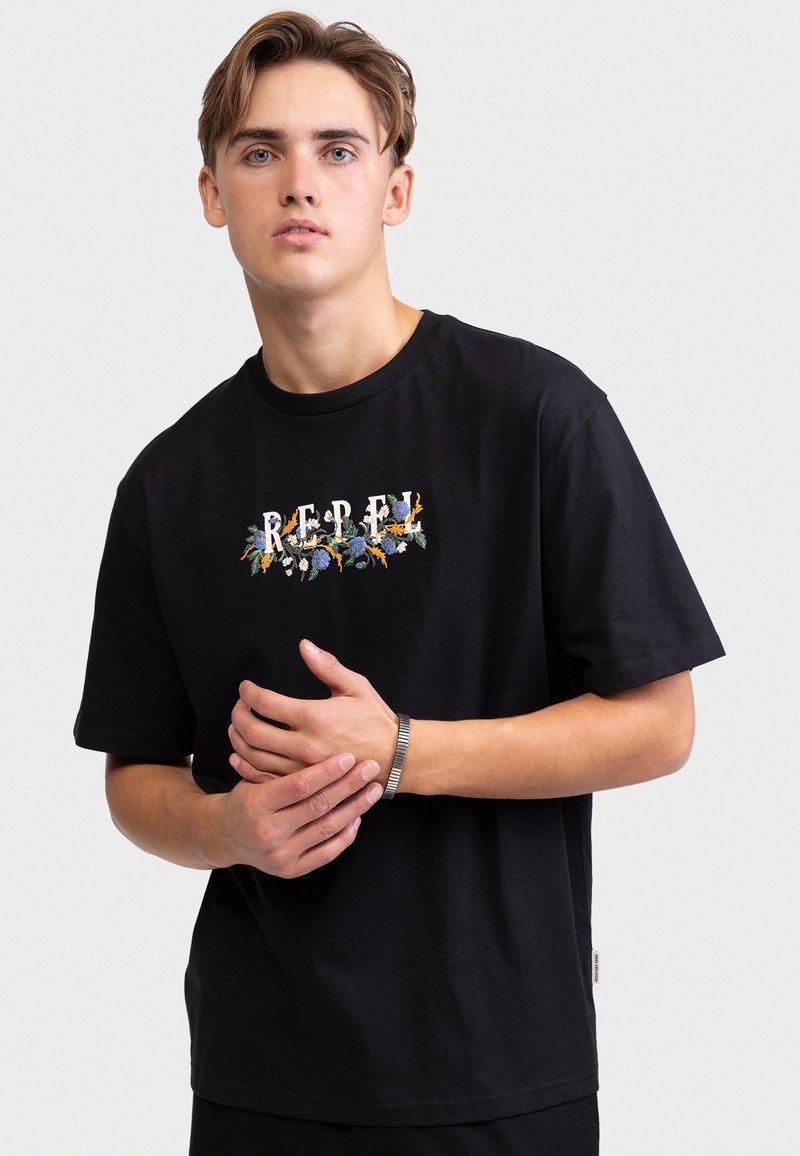 Νέος άνδρας που φοράει μαύρο t-shirt με τη λέξη "REBEL" διακοσμημένη με πολύχρωμα λουλουδένια σχέδια και ασημένιο βραχιόλι στον καρπό του.