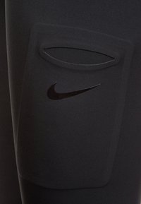 Musta urheilulliset leggingsit, joissa on sivutasku kaarevalla aukolla ja musta Nike-logo taskun yläpuolella. Sileä, joustava kangas.