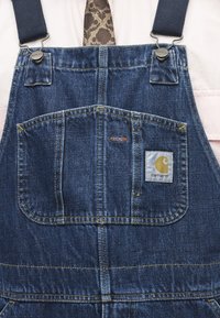 Peto de mezclilla Carhartt con bolsillo frontal y parche con el logo, combinado con una camisa rosa claro y una corbata marrón con estampado.