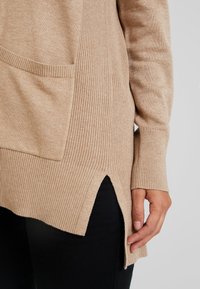 Pull en maille beige avec une fente latérale, ourlet côtelé, manches longues, poche avant, porté sur un pantalon noir avec une main visible sur le côté.