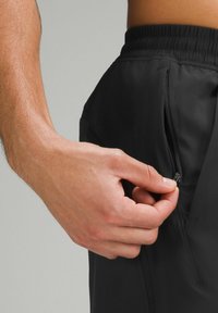 Black athletic shorts with a zippered pocket, featuring a smooth texture and elastic waistband. A hand grips the zipper for access.